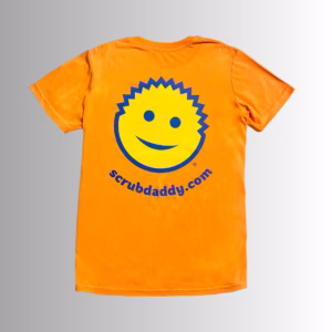 Image Description: A promotional product image showing the back of the vibrant orange short-sleeve t-shirt set against a neutral gradient background, featuring a large yellow smiley-face graphic with blue outlines and facial features above the blue URL text "scrubdaddy.com"