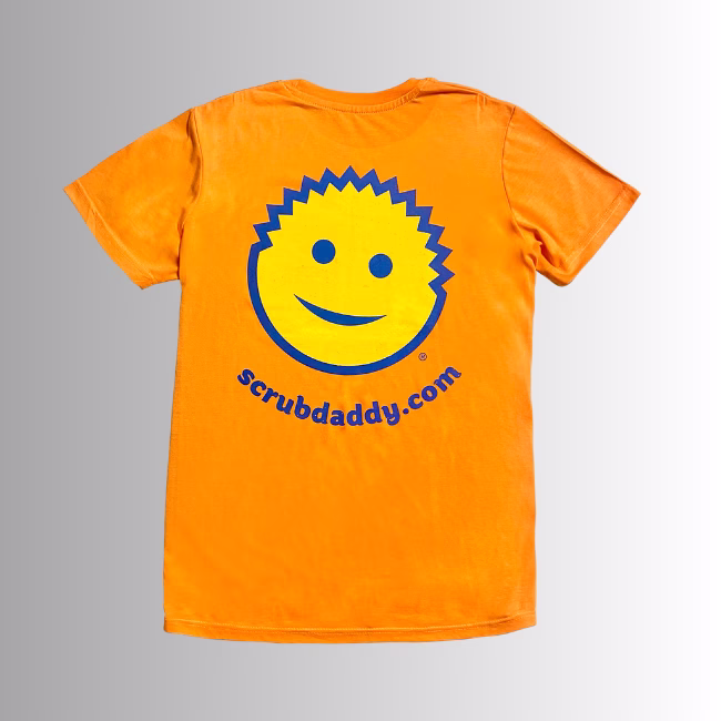 Image Description: A promotional product image showing the back of the vibrant orange short-sleeve t-shirt set against a neutral gradient background, featuring a large yellow smiley-face graphic with blue outlines and facial features above the blue URL text "scrubdaddy.com"