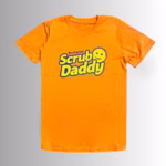 Image Description: A promotional product hero image of the front of a vibrant orange short-sleeve t-shirt set against a neutral gradient background, featuring the blue and yellow "The Original Scrub Daddy" logo printed across the chest.