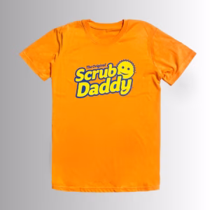 Image Description: A promotional product hero image of the front of a vibrant orange short-sleeve t-shirt set against a neutral gradient background, featuring the blue and yellow "The Original Scrub Daddy" logo printed across the chest.