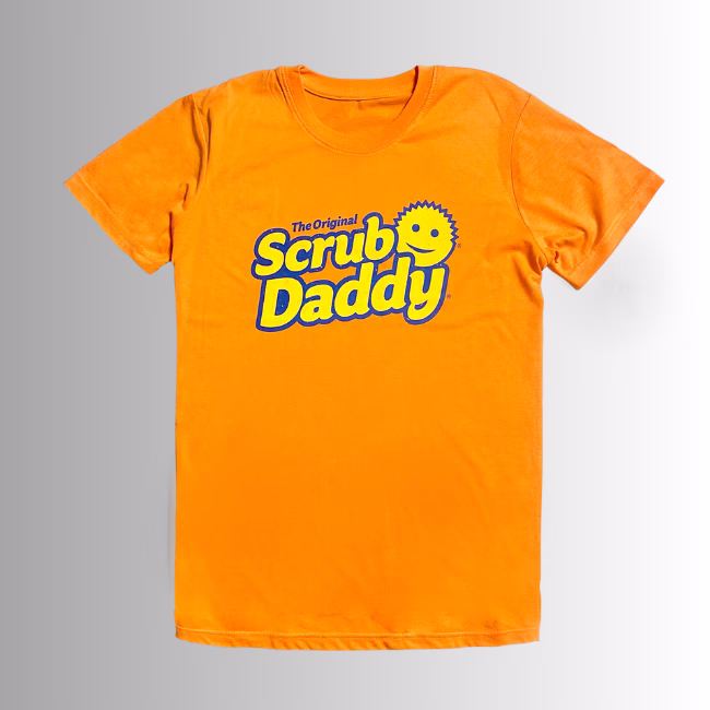 Orange t-shirt front Image Description: A promotional product hero image of the front of a vibrant orange short-sleeve t-shirt set against a neutral gradient background, featuring the blue and yellow "The Original Scrub Daddy" logo printed across the chest.