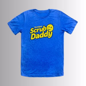 Image Description: A promotional product hero image of the front of a royal blue short-sleeve t-shirt set against a neutral gradient background, featuring the yellow "The Original Scrub Daddy" logo printed across the chest.
