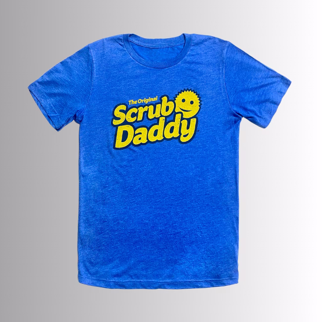 Image Description: A promotional product hero image of the front of a royal blue short-sleeve t-shirt set against a neutral gradient background, featuring the yellow "The Original Scrub Daddy" logo printed across the chest.