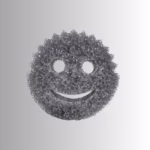 Image Description: A promotional product hero image of a classic smiley-face scrubbing sponge in a textured metallic dark grey or charcoal finish, set against a neutral gradient background.