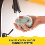 Image Description: A lifestyle product photo featuring a pair of hands rinsing a grey textured smiley-face sponge under a kitchen faucet with running water, accompanied by yellow text at the bottom that reads "Rinses Clean Under Running Water".