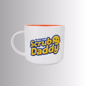 Image Description: A promotional product hero image of a white ceramic 16oz coffee mug set against a neutral gradient background, featuring a comfortable side handle, a vibrant orange interior glaze, and "The Original Scrub Daddy" logo printed on the outer side.