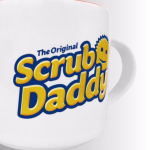 Image Description: A close-up product detail shot of the white ceramic mug set against a neutral gradient background, emphasizing the glossy print quality of "The Original Scrub Daddy" logo with its iconic smiling face icon.