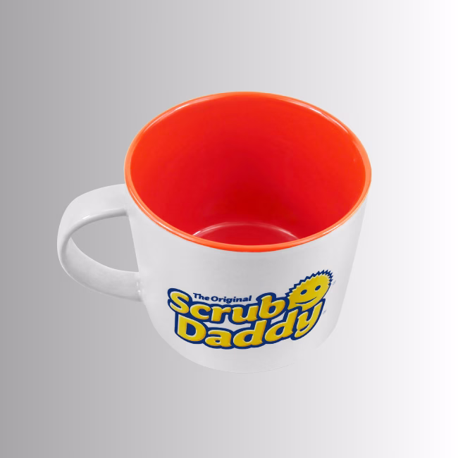 Image Description: A high-angle product shot of the white ceramic Scrub Daddy 16oz mug set against a neutral gradient background, showcasing its bright orange inner lining and the side logo.