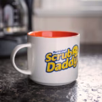 Image Description: A lifestyle product photo of the white and orange Scrub Daddy 16oz mug sitting on a dark stone kitchen countertop next to an appliance.