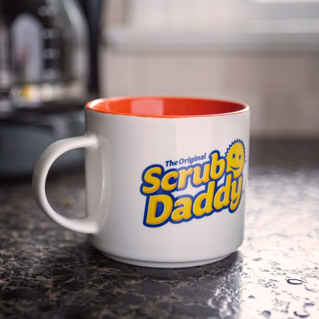 Image Description: A lifestyle product photo of the white and orange Scrub Daddy 16oz mug sitting on a dark stone kitchen countertop next to an appliance.