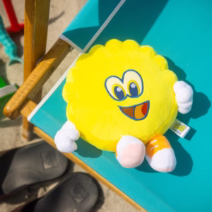 mage Description: A lifestyle product photo of the yellow Scrub Daddy plush toy resting on the turquoise fabric of a beach chair next to a pair of sandals and sand toys.