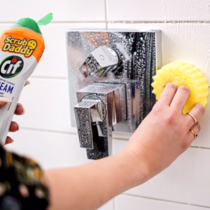 Image Description: A lifestyle action shot of a person using a yellow Scrub Daddy sponge in one hand to scrub white tiled walls next to a chrome shower handle, while holding a bottle of Scrub Daddy Cif Cream cleanser in the other hand.