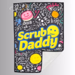 Image Description: A promotional product hero image of a black Scrub Daddy golf towel set against a neutral gradient background, featuring an all-over doodled pattern with colorful smiley faces and geometric designs, large yellow "Scrub Daddy" lettering across the center, a silver carabiner clip in the upper corner, and a folded corner revealing the white backing.