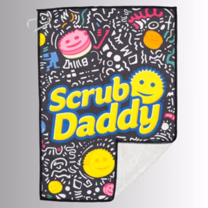 Image Description: A promotional product hero image of a black Scrub Daddy golf towel set against a neutral gradient background, featuring an all-over doodled pattern with colorful smiley faces and geometric designs, large yellow "Scrub Daddy" lettering across the center, a silver carabiner clip in the upper corner, and a folded corner revealing the white backing.