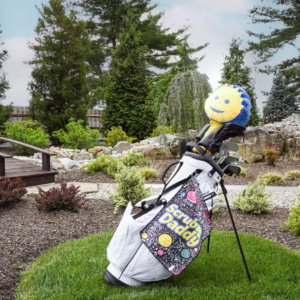 Image Description: An outdoor lifestyle action shot of a white golf bag standing on a manicured lawn near a garden path and bridge, featuring the black Scrub Daddy doodled golf towel clipped to the bag alongside a blue-and-yellow Sponge Daddy club cover.