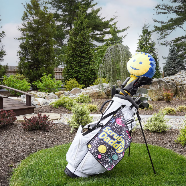 Image Description: An outdoor lifestyle action shot of a white golf bag standing on a manicured lawn near a garden path and bridge, featuring the black Scrub Daddy doodled golf towel clipped to the bag alongside a blue-and-yellow Sponge Daddy club cover.