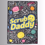 Image Description: A promotional product flat-lay image of a black Scrub Daddy golf towel against a neutral gradient background, featuring an all-over doodled pattern with colorful smiley faces, geometric shapes, and large yellow "Scrub Daddy" lettering across the center.