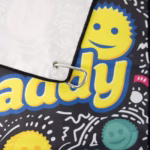 Image Description: A close-up product detail shot focusing on the corner of the black Scrub Daddy golf towel, highlighting the silver carabiner clip attached through a metal grommet and the vibrant yellow "Scrub Daddy" typography against the patterned background.