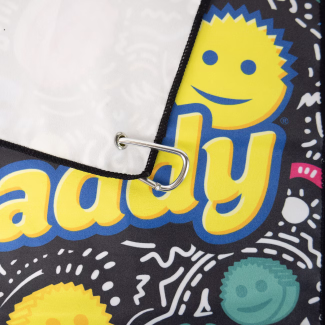Image Description: A close-up product detail shot focusing on the corner of the black Scrub Daddy golf towel, highlighting the silver carabiner clip attached through a metal grommet and the vibrant yellow "Scrub Daddy" typography against the patterned background.