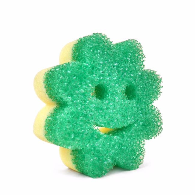A green flower-shaped Scrub Mommy sponge shown from an angled perspective against a solid white background, highlighting its dual-sided design with a textured green FlexTexture scrubbing side on the front and a yellow absorbent foam side visible along the edges.
