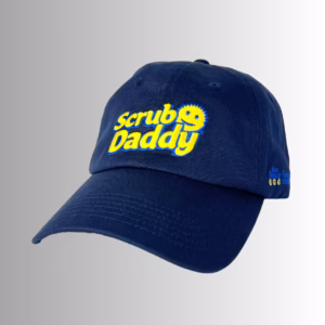 Image Description: A promotional product hero image of a navy blue Scrub Daddy baseball cap set against a neutral gradient background, featuring bold embroidered yellow and blue "Scrub Daddy" lettering on the front crown and detailed side embroidery.