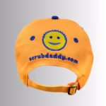 Image Description: A promotional product image showing the back of the orange Scrub Daddy baseball cap set against a neutral gradient background, highlighting an embroidered yellow and blue smiley-face logo above the URL "scrubdaddy.com" and an adjustable fabric strap with a metal buckle.