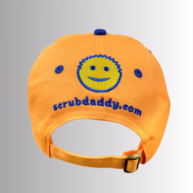 Image Description: A promotional product image showing the back of the orange Scrub Daddy baseball cap set against a neutral gradient background, highlighting an embroidered yellow and blue smiley-face logo above the URL "scrubdaddy.com" and an adjustable fabric strap with a metal buckle.
