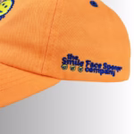 Image Description: A close-up product detail shot of the side of the orange Scrub Daddy baseball cap set against a neutral gradient background, showcasing blue embroidered text that reads "the Smile Face Sponge company" alongside small smiley face icons.