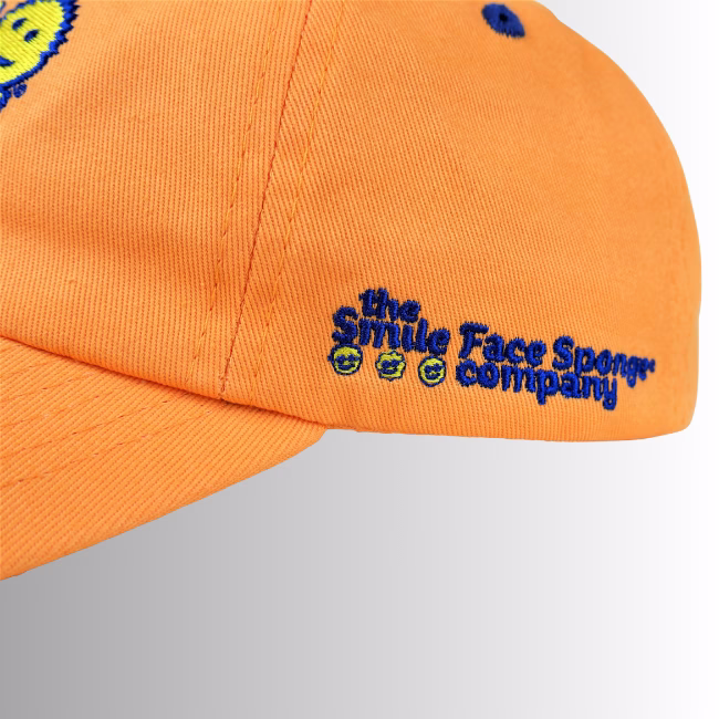 Image Description: A close-up product detail shot of the side of the orange Scrub Daddy baseball cap set against a neutral gradient background, showcasing blue embroidered text that reads "the Smile Face Sponge company" alongside small smiley face icons.