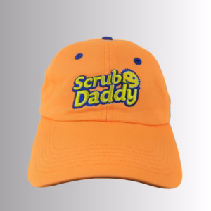Image Description: A promotional product hero image of an orange Scrub Daddy baseball cap set against a neutral gradient background, featuring embroidered blue and yellow "Scrub Daddy" lettering on the front crown.