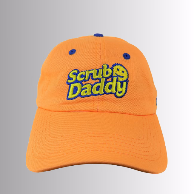Scrub Daddy Baseball Cap Orange hero Image Description: A promotional product hero image of an orange Scrub Daddy baseball cap set against a neutral gradient background, featuring embroidered blue and yellow "Scrub Daddy" lettering on the front crown.