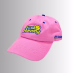 Image Description: A promotional product hero image of a pink Scrub Mommy baseball cap set against a neutral gradient background, featuring embroidered blue and yellow "Scrub Mommy" lettering with a small bow icon on the front crown, and side embroidery reading "the Smile Face Sponge company".
