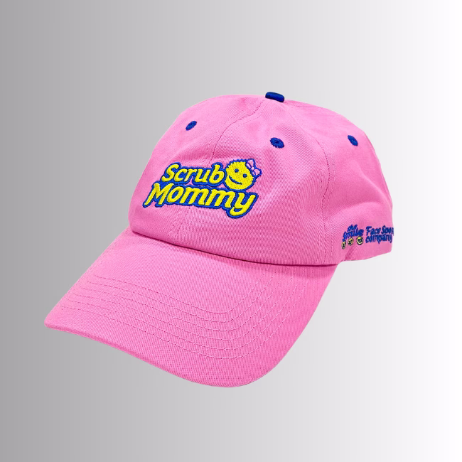 Image Description: A promotional product hero image of a pink Scrub Mommy baseball cap set against a neutral gradient background, featuring embroidered blue and yellow "Scrub Mommy" lettering with a small bow icon on the front crown, and side embroidery reading "the Smile Face Sponge company".