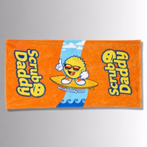 Image Description: A promotional product flat-lay image of a vibrant orange beach towel set against a neutral gradient background, featuring a central graphic of a smiling Scrub Daddy character wearing sunglasses and sneakers while riding a surfboard on a blue wave path, flanked by large blue and yellow "Scrub Daddy" lettering on either side.