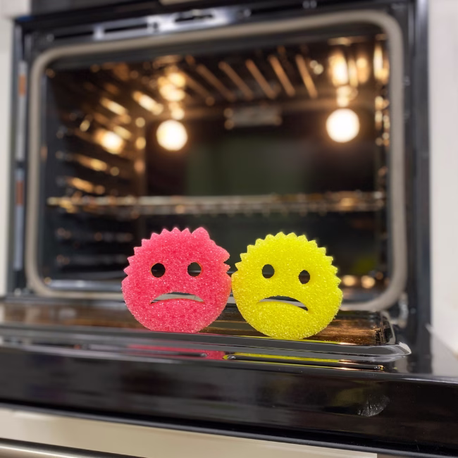 Image Description: A lifestyle product photo featuring a pink and a yellow Cranky Couple sponge resting on the open door frame of a clean kitchen oven with its internal lights illuminated.