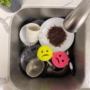 Image Description: A kitchen sink lifestyle action shot featuring a yellow and a pink Cranky Couple sponge sitting among dirty dishes, including a plate with baked-on residue, a mug, and glasses in a stainless steel sink next to a faucet.