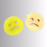 Image Description: A promotional product flat-lay image set against a neutral gradient background, featuring one yellow and one pink Cranky Couple scrubbing sponge with textured edges and frowning facial expressions.