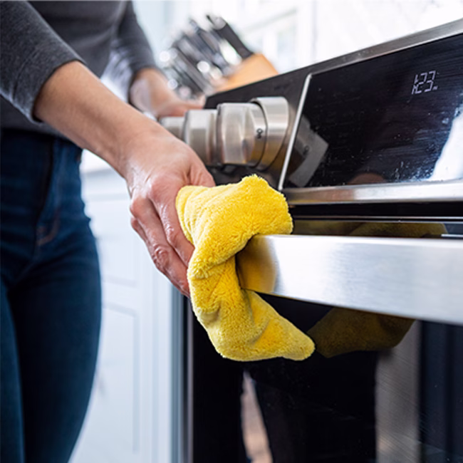 Image Description: A close-up shot of a person's hand using a bright yellow microfiber cloth to wipe down the stainless steel handle of a modern oven range in a kitchen setting.