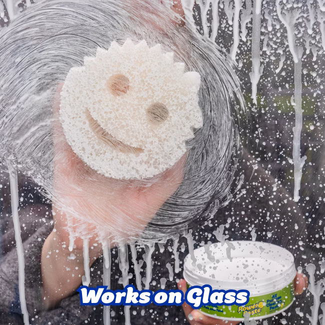 Image Description: A lifestyle action shot showing a hand using a Scrub Mommy sponge with soapy water and PowerPaste to clean and scrub smudges off a glass window or surface, with an open tub of PowerPaste visible in the corner.