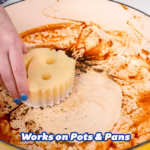 Image Description: A lifestyle action shot of a hand using a dual-sided Scrub Mommy sponge with PowerPaste to scrub away baked-on grease and food residue inside a stained pot or pan.