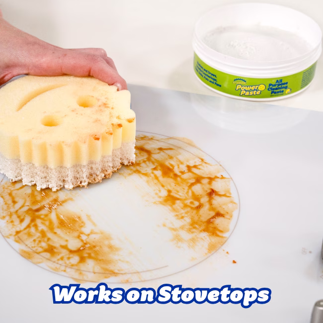 Image Description: A lifestyle action shot of a hand using a dual-sided Scrub Mommy sponge to clean a burnt-on spill and brown residue off a white glass stovetop, with an open tub of PowerPaste resting nearby.