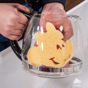 A close-up shot of hands holding a yellow, soft-textured side of a Minnie Mouse-shaped Scrub Mommy sponge inside a glass coffee pot to clean it. © Disney