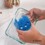 Image Description: Scrub Daddy Disney Stitch Sponges (3-Pack) being used to wash a glass baking dish under a running kitchen faucet. Hands hold the blue Stitch character sponge inside the dish as water streams over it. The copyright "© Disney" is displayed in the bottom right corner.