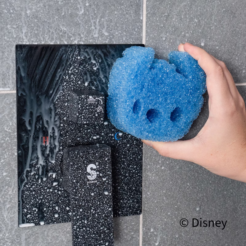 Image Description: Scrub Daddy Disney Stitch Sponges (3-Pack) being used for bathroom cleaning. A hand holds the blue Stitch character sponge next to a soapy, textured shower fixture and tiled wall. The copyright "© Disney" is displayed in the bottom right corner.