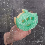 Image Description: A hand holding a green Alien-shaped Scrub Mommy sponge covered in soap bubbles against a wet, soapy glass shower door background. © Disney/Pixar