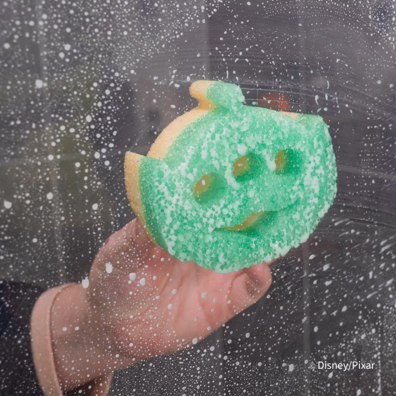 Image Description: A hand holding a green Alien-shaped Scrub Mommy sponge covered in soap bubbles against a wet, soapy glass shower door background. © Disney/Pixar
