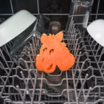 Image Description: An orange Bullseye-shaped Scrub Mommy sponge placed upright on the bottom wire rack of a dishwasher, surrounded by glassware and dishes in a kitchen setting. © Disney/Pixar