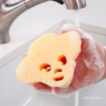 Image Description: A close-up shot of a hand holding a yellow and red Jessie-shaped Scrub Mommy sponge covered in soapy lather, being rinsed under a running kitchen faucet against a clean white backdrop. © Disney/Pixar