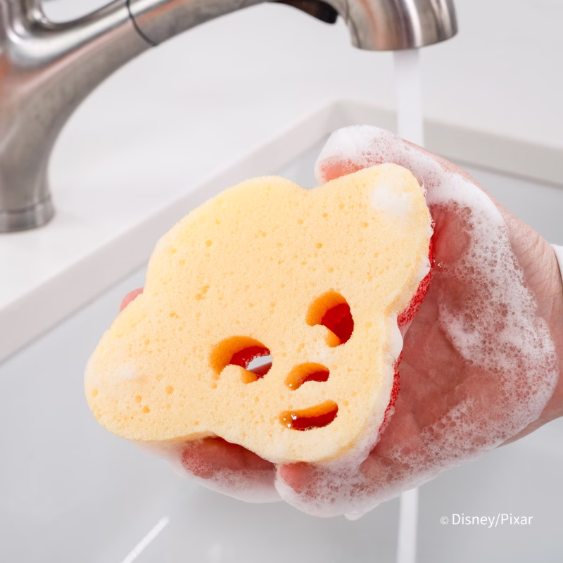 Image Description: A close-up shot of a hand holding a yellow and red Jessie-shaped Scrub Mommy sponge covered in soapy lather, being rinsed under a running kitchen faucet against a clean white backdrop. © Disney/Pixar