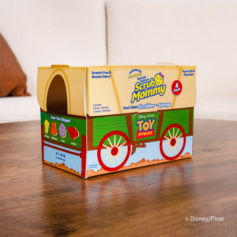 Image Description: A product display of the Toy Story Scrub Mommy 4-count collector's edition packaging, designed to look like a red and green western wagon toy chest sitting on a wooden surface in front of a neutral background. © Disney/Pixar